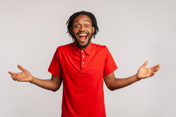 哇太棒了!长着骇人辫、身穿红色休闲风格t恤的迷人男子，惊讶地站在那里，双臂高举，嘴巴张得大大的。图片下载