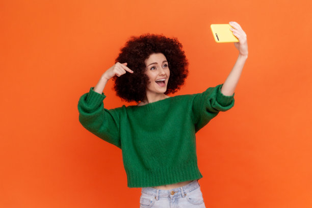 年轻的成年女性，卷发，穿着绿色休闲风格的毛衣直播，手指向下，订阅。图片下载