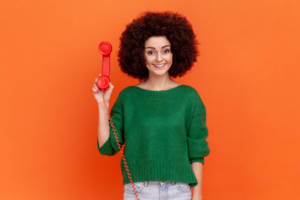 照片上的女人微笑着卷发穿着绿色休闲风格的毛衣站着拿着红色复古的固定电话，等待交谈。图片下载