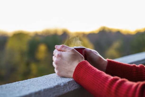 女子端着一杯热气腾腾的饮料，站在高楼阳台的边缘，背景是模糊的树木和夕阳图片下载
