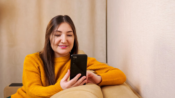 穿着黄色毛衣的年轻女子坐在棕色沙发上看短信图片下载
