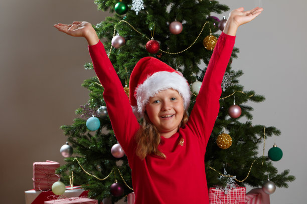 快乐的小女孩戴着圣诞老人的帽子。那孩子在圣诞树的背景下举起双手。图片下载