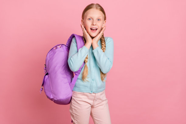 可爱的印象深刻的学校女孩穿着蓝色开襟羊毛衫背包手臂脸颊微笑孤立的粉红色背景图片下载