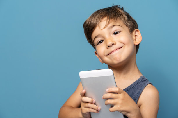正面照片中四岁的白人小男孩站在蓝色背景摄影棚前，站着用数码平板看着相机开心地笑着图片下载