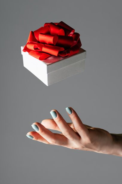 女性的手扔到空中或抓住白色的礼物盒与红色的蝴蝶结在灰色的背景。悬浮的礼物,垂直素材图片