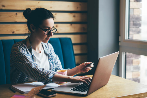 年轻女子戴着眼镜，挽着发髻，在自助餐厅里，坐在带着笔记本电脑的木桌前，从网站折扣卡输入promocode。图片下载