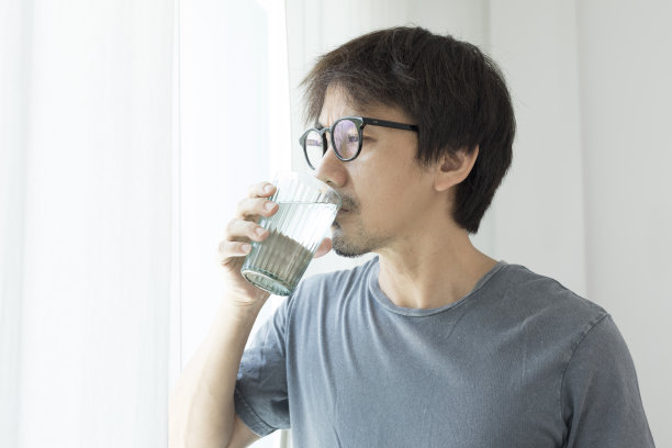 一个亚洲人站在家里的窗户边喝水。图片下载