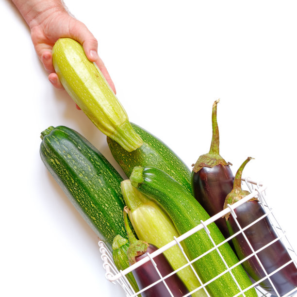 用蔬菜成分。整个成熟的西葫芦和茄子放在白色背景的金属柳条篮子里。一个男人的手从一包蔬菜中取出一份。秋天,秋天的概念。收割。食品配料。平的。图片下载