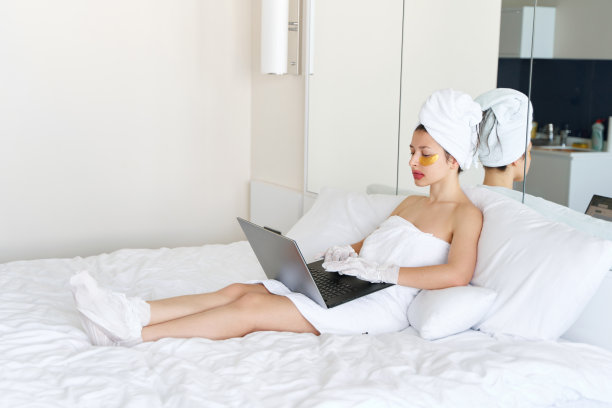 洗完澡后，一个女孩裹着毛巾在笔记本电脑前工作，用化妆品贴在眼睛、嘴唇和手套下面的皮肤上，为她的手脚保湿。家用身体护理化妆品趋势图片下载
