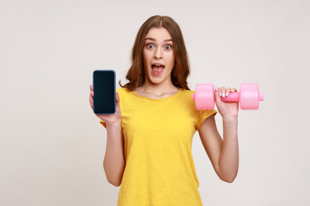 一个年轻的女人穿着黄色的t恤，手里拿着空屏幕的智能手机和粉红色的哑铃，为新的健身应用做广告。图片下载