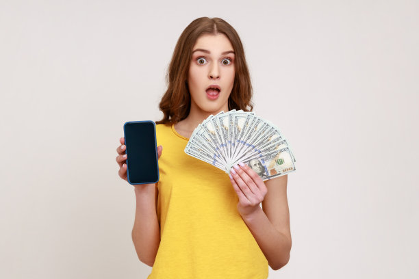 哇,有利可图的服务。照片中的少女身穿黄色T恤，手里拿着美元和智能手机，在手机应用中赚钱。图片下载