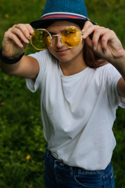 离焦特写一个快乐的棕色头发的年轻女人戴着一顶帽子在户外。显示黄色太阳镜。绿色的自然背景。看相机。垂直的。的焦点图片下载