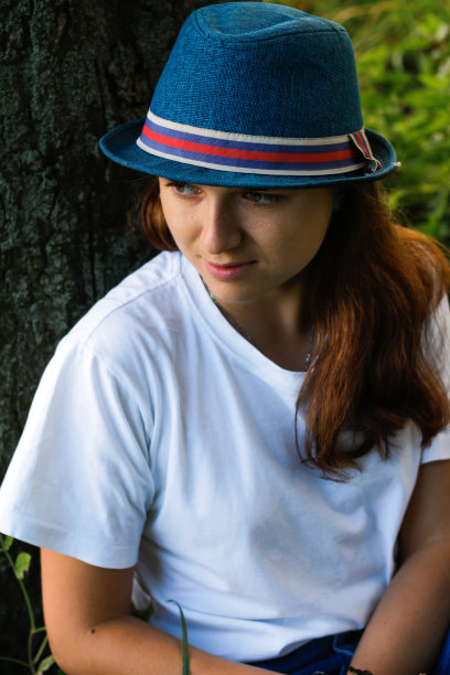 离焦特写，一个浪漫梦幻的年轻女人，棕色头发，戴着帽子，白色t恤在户外。你好,秋天。女人坐在树干旁。的焦点图片下载