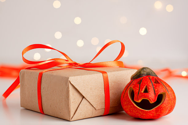 特写的棕色礼盒与橙色缎带蝴蝶结和小微笑万圣节南瓜明亮模糊的背景与bokeh复制空间。秋天节日的概念图片下载