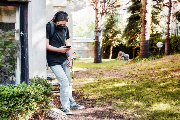 年轻的高中男孩带着医用口罩、耳机和背包，用他的智能手机发短信图片下载