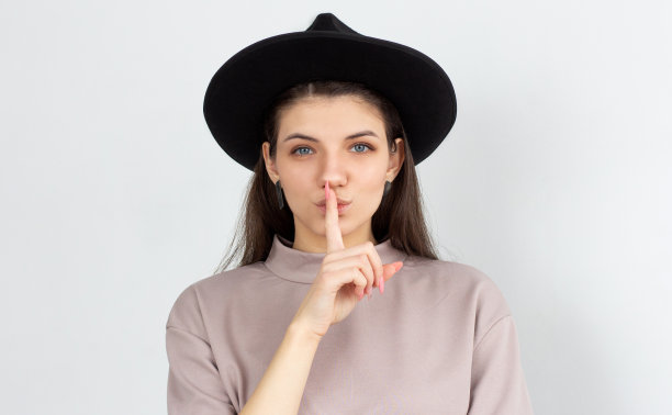 女人准备告诉美丽的秘密。迷人的轻佻的白人女性肖像作出嘘或嘘的手势与食指在嘴，看起来性感和自信的相机在白色的墙图片下载