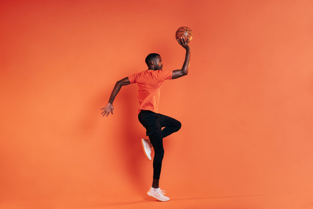 运动员手拿篮球在空中跳跃的侧面。穿着运动服的男子在室内橘色的墙壁前锻炼。图片下载