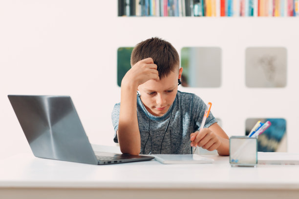 小男孩戴着耳机坐在桌子旁，拿着笔记本电脑，准备上学。在线教育的概念。图片下载