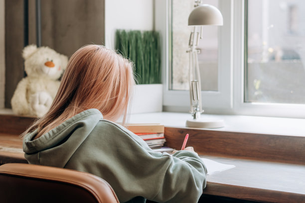 漂亮的金发少女在家里的衣服做作业，在她的书桌上做笔记，背面。学校的概念图片下载