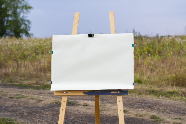 在乡村风景的背景下，一个艺术画架上放着一张空白的画纸。图片下载