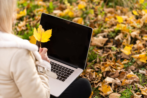 秋天公园里带着笔记本电脑的美女。美丽的自然景色与多彩的树叶背景，黄色的树木和树叶在秋天。秋季户外生活方式。落叶上快乐微笑的女人图片下载