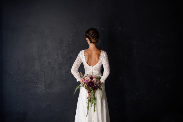 时尚新娘身穿白色镂空婚纱，无头纱，背站着，手捧婚礼花束图片下载