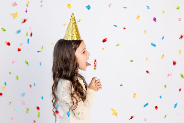 一个可爱的小女孩正在吃一个大棒棒糖，她戴着节日帽，背景是白色的，背景上有一个飘带。空间的文本。小女孩庆祝生日，节日概念图片下载