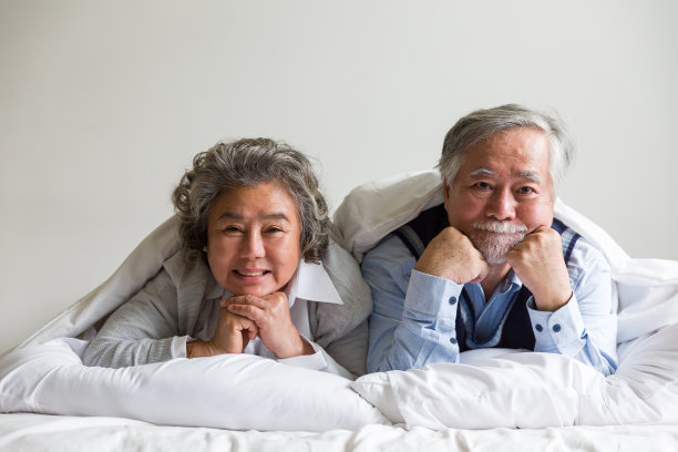 幸福的老夫妇躺在床上，床上有白色的毯子，枕头在家里的卧室里。退休、医疗保健、放松和花时间的概念图片下载