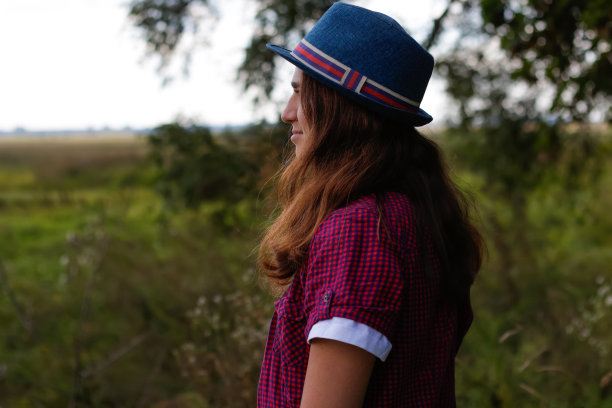 离焦近距离拍摄一名微笑的年轻女子，棕色头发，戴着一顶帽子在户外。绿色的自然背景。后视图。的焦点图片下载