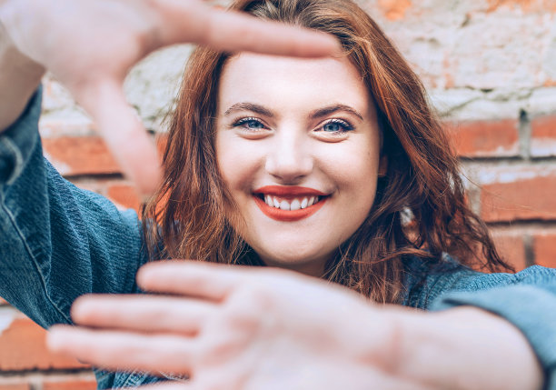 真诚微笑的肖像红色卷曲的长发白种人妇女应用红色口红的嘴唇，蓝色的眼睛，红色砖墙背景做一个“没有照片”的手势。图片下载