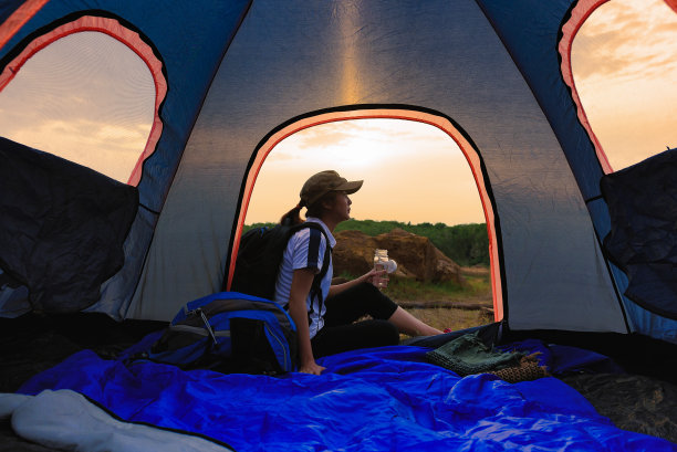 年轻的女旅行者穿着运动服，坐在蓝色的帐篷里喝水，在日落时休息在大自然中，透过帐篷看。游客户外露营，背包旅行。图片下载