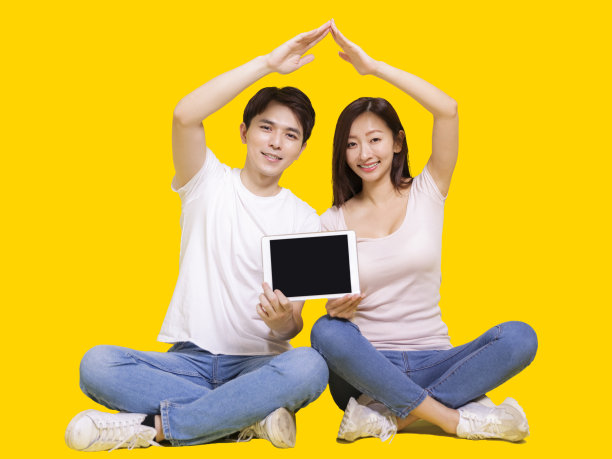 年轻幸福的夫妇与屋顶房子的手势和平板。孤立在黄色背景上。图片下载