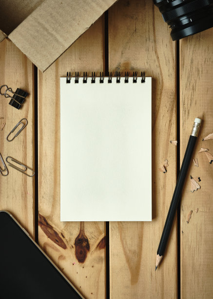 顶部表格视图，打开的空白笔记本在木制书桌上与打开的硬纸板盒，相机镜头，铅笔，手机，平铺的文本复制空间。图片下载