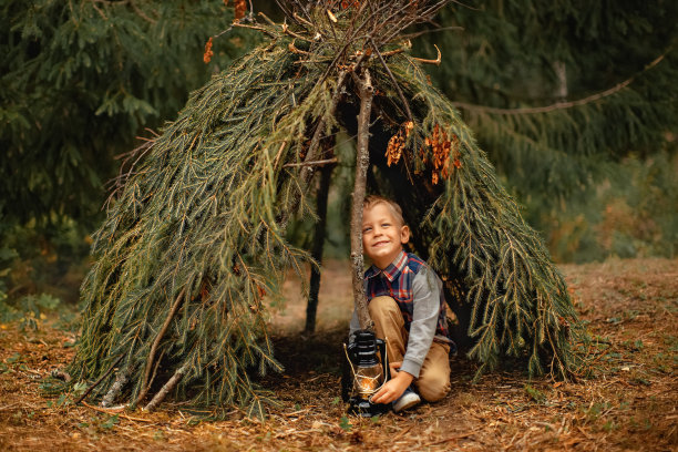 夏天的森林里，一个小男孩坐在手工搭建的小屋里，手里拿着一盏灯图片下载