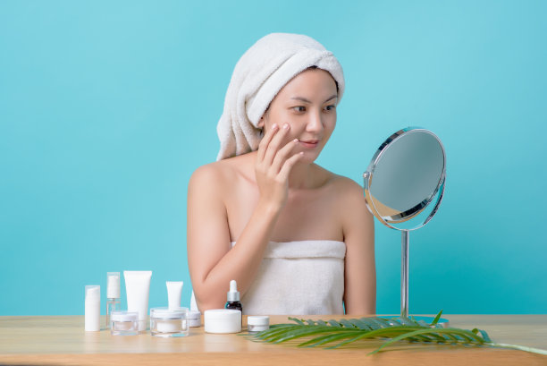 美丽的亚洲年轻女子微笑着触摸脸，看着镜子与木制桌子上的化妆品模型。护肤清洁理念，返老还童的美容治疗过程。图片下载