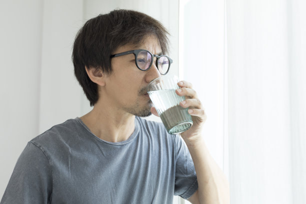 一个亚洲人站在家里的窗户边喝水。图片下载