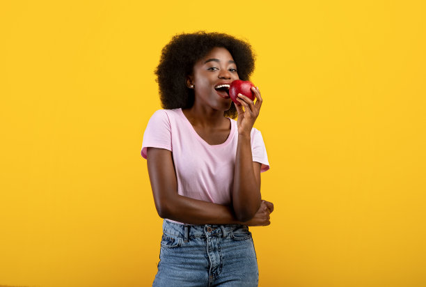 年轻的非洲裔美国女士带着美丽的微笑吃着美味的红苹果，宣传健康营养，黄色背景图片下载