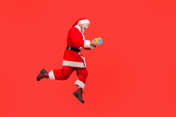 满脸灰胡子的老人穿着圣诞老人的服装心满意足地过着寒假，拿着包装好的礼盒高高跳着。图片下载