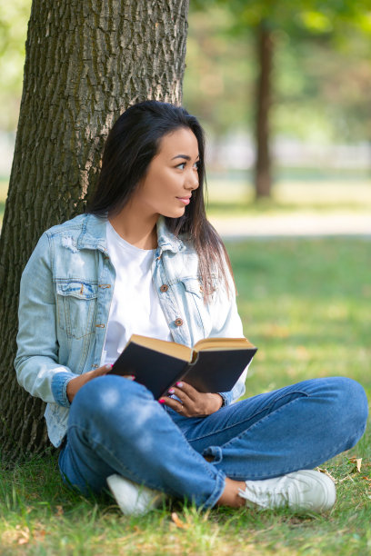 一个亚洲女人在公园里拿着一本书的垂直照片。她坐在公园的草地上，并排看着。休息的概念来自电子产品、智能手机和互联网图片下载