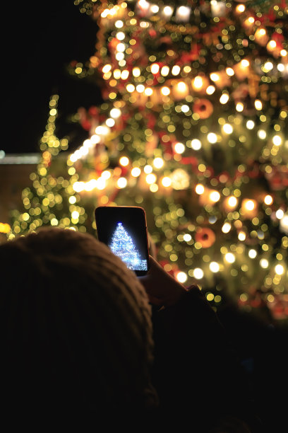 一个人用智能手机拍下一棵装饰精美的圣诞树。节日，圣诞节庆祝活动，节假日。艺术模糊,散景。图片下载