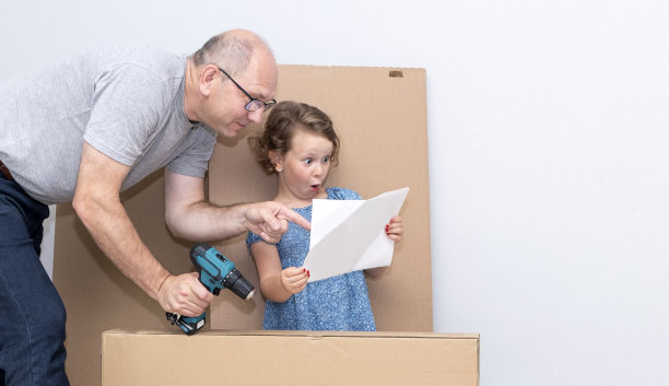 一个六岁的女孩和一个白人男子。一家人看了看大纸板箱的背景。他们看了家具组装计划。那孩子收到了一份礼物，育儿室的家具。图片下载