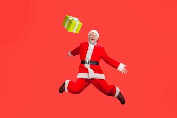 穿着圣诞老人服装的满脸灰胡子的老人跳得很高，把礼物抛在空中，双手摊开。图片下载