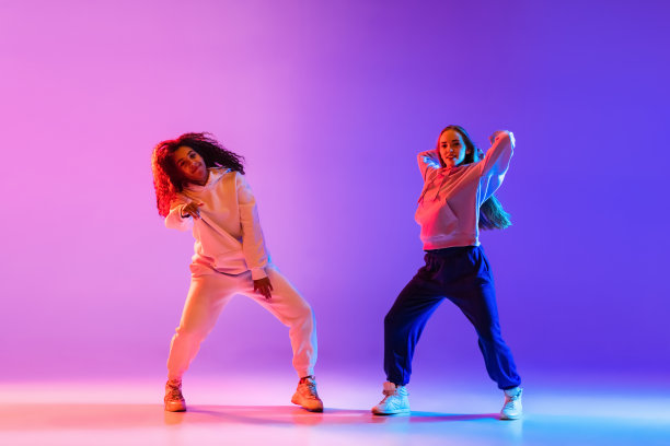 两个美丽的年轻女孩在霓虹灯的彩色渐变背景下穿着时尚的衣服跳街舞。图片下载
