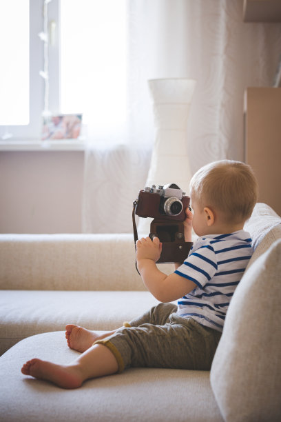 可爱的男孩在玩古董相机。一个孩子坐在客厅的沙发上，手里拿着相机想要自拍。儿童早期发展。色调和人工噪音图片下载