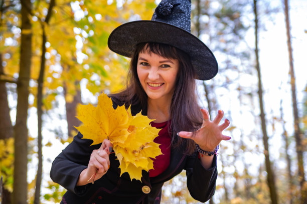 微笑的女巫戴着黑色的大帽子庆祝万圣节，女人手持金色的枫叶在秋天的森林背景上图片下载