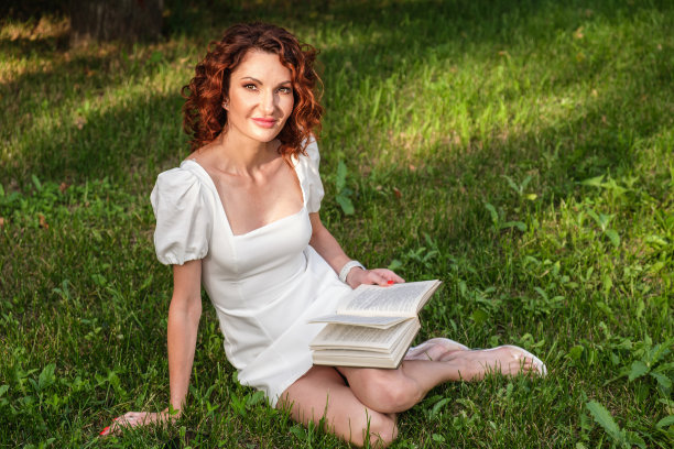 漂亮苗条的年轻女子，一头卷发，身穿白色连衣裙，手里拿着一本书，坐在草坪上图片下载