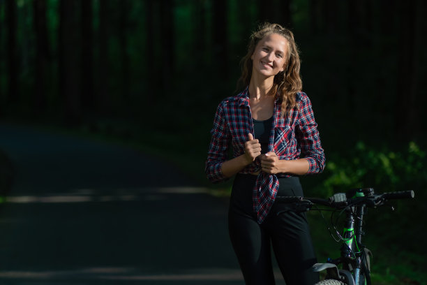 在一个阳光明媚的夏日，一个穿着衬衫和打底裤的微笑的年轻女人在公园里骑着自行车，复制空间图片下载