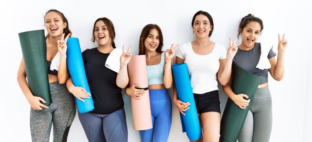 一群妇女拿着瑜伽垫，站在孤立的背景下，微笑着笑脸，对着镜头眨眼，用手指做胜利的手势。二号人物。图片下载