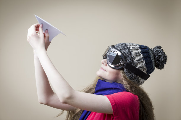 微笑的白种人金发少女在飞行眼镜手持折纸飞机在米色背景。水平的形象定位图片下载