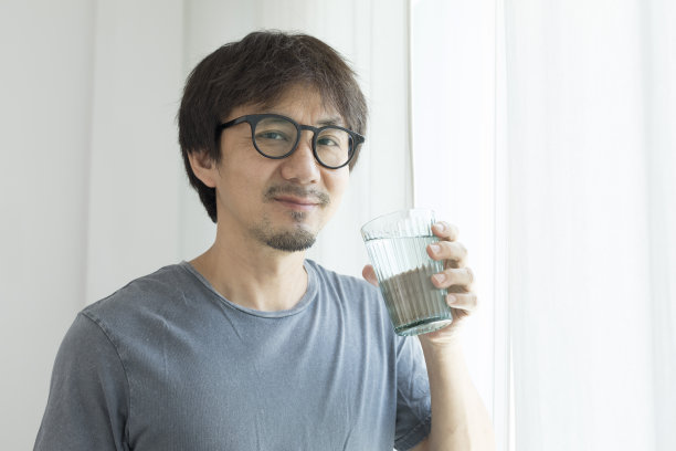 一个亚洲人站在家里的窗户边喝水。图片下载